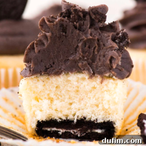 An oreo bottom cupcake with vanilla cake, a chocolate cookie at the base, and thick chocolate frosting on top. The cupcake sits in a white paper liner with a bite taken out, revealing its delicious layers.