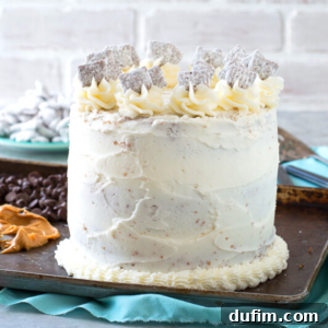 A tall, round puppy chow cake with white frosting, decorated with swirls of icing and powdered sugar-covered animal cookies on top, sits on a metal tray with chocolate chips and cookies in the background.