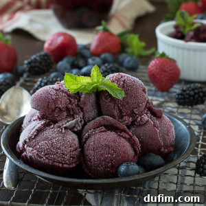 A bowl of sangria sorbet garnished with a mint leaf, surrounded by fresh strawberries, blueberries, and blackberries on a cooling rack, with a spoon nearby.