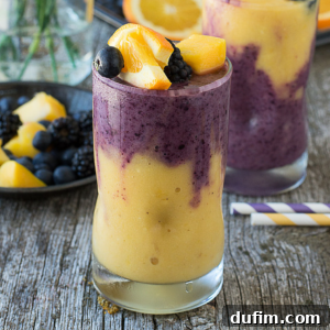 Two glasses of Hawaiian Berry Smoothie, with yellow and purple swirls, are topped with peach slices, blackberries, and blueberries on a rustic wooden surface. A striped straw and plate of fruit complete the vibrant scene.