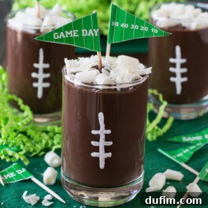 Three football pudding cups are topped with white icing laces and white chocolate curls, each adorned with a green Game Day flag. Green shredded paper decorates the festive background.