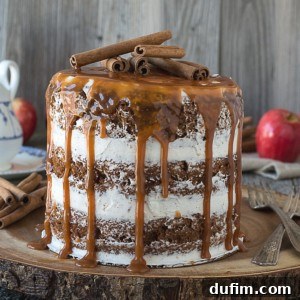 Harvest Apple Spice Cake with Golden Caramel Drizzle 7 A tall apple spice cake with white frosting between layers is topped with dripping caramel sauce and several cinnamon sticks, sitting on a wooden board. Red apples and utensils are visible in the background.