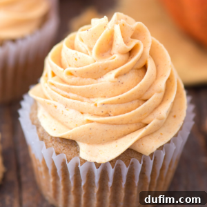 A close-up of a cupcake with light orange, pumpkin buttercream swirled frosting, set in a white paper liner on a wooden surface. The frosting appears creamy and lightly spiced.