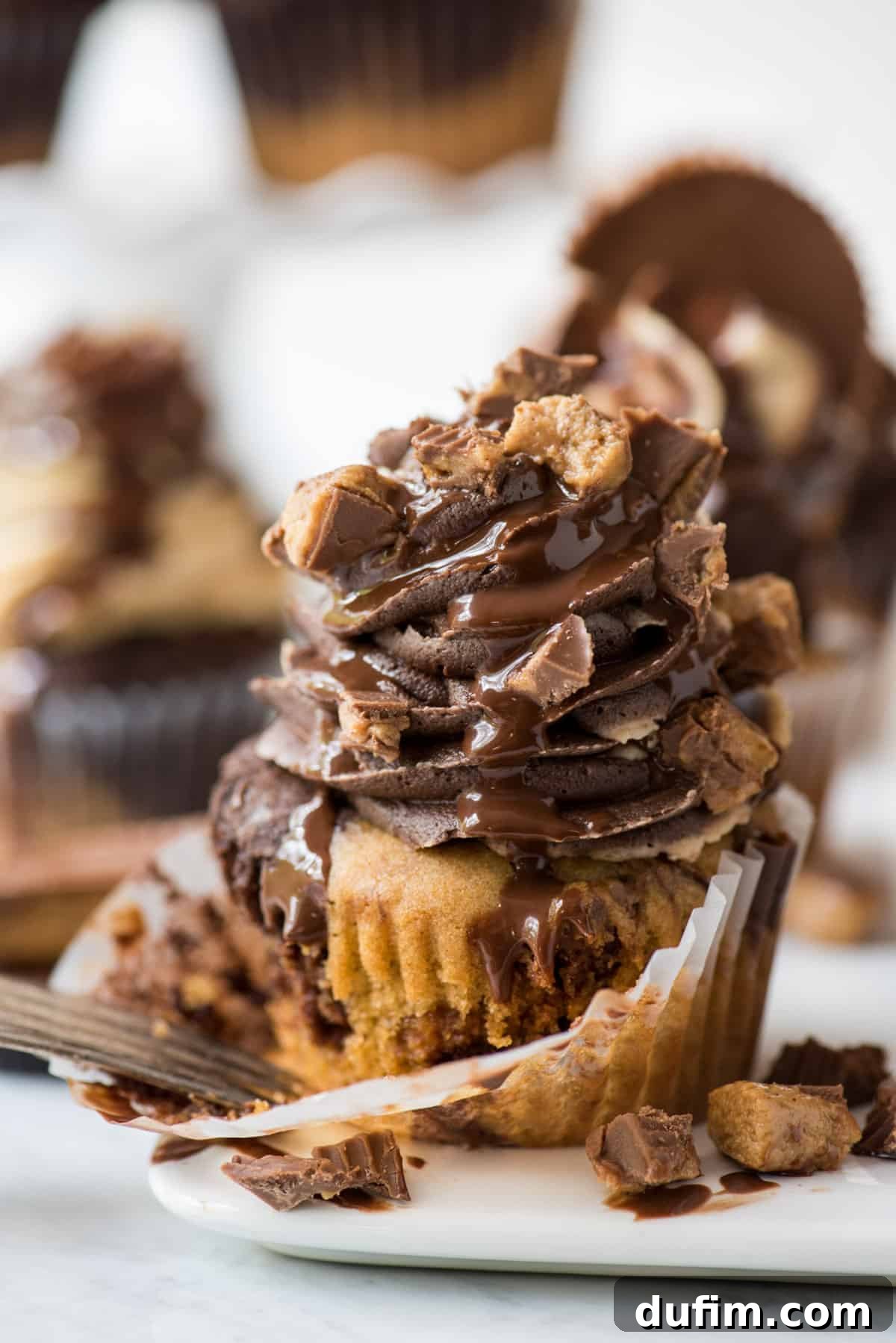 chocolate peanut butter cupcake with swirled chocolate peanut butter frosting topped with chopped reese’s and drizzled with chocolate sauce on white background