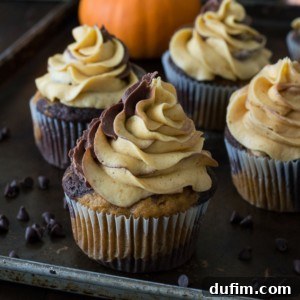 A close-up of pumpkin chocolate cupcakes and vanilla swirled cupcakes with creamy, two-toned frosting on top, sitting on a baking tray. Chocolate chips are scattered around, and a small pumpkin is in the background.