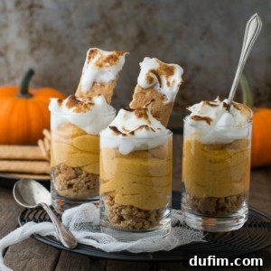 Three glasses filled with layered pumpkin mousse, crumbled graham crackers, and topped with whipped cream recreate festive pumpkin pie shooters. Two whole pumpkins and extra crackers are in the background on a rustic wooden table.