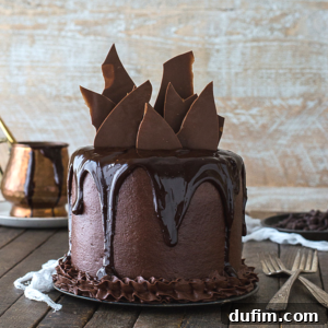 A tall chocolate chocolate cake with glossy ganache dripping down the sides, topped with large shards of chocolate, sits on a wooden table with forks and a copper pot in the background.