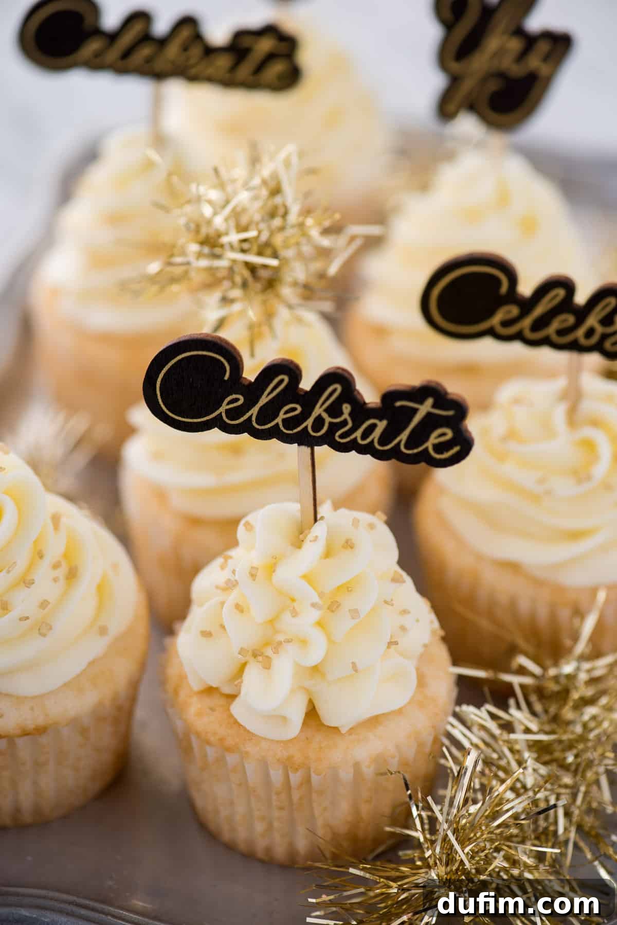 Another view of easy champagne cupcakes, frosted with creamy champagne buttercream and adorned with gold 'Celebrate' toppers, glittering sprinkles, and gold tinsel, perfect for New Year's festivities.