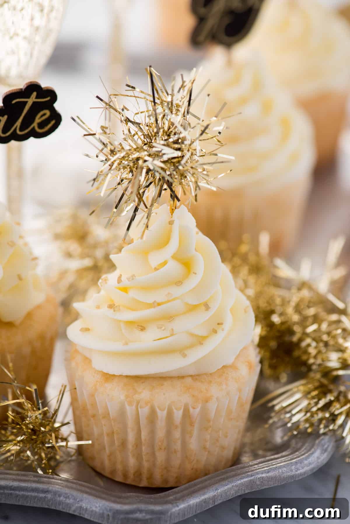 A close-up of a single easy champagne cupcake, generously frosted with champagne buttercream, adorned with sparkling gold sprinkles and a festive gold tinsel cupcake topper.