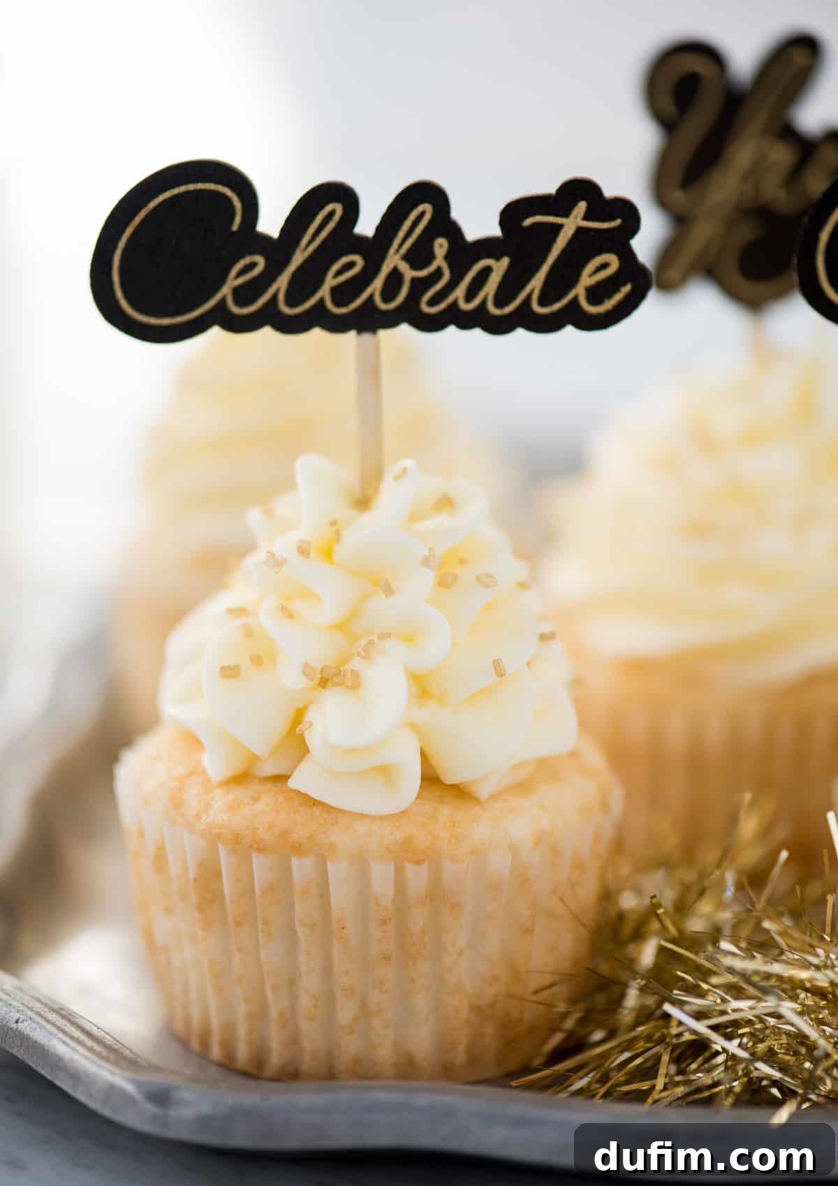 A selection of easy champagne cupcakes frosted with champagne buttercream, topped with various gold sprinkles and 'Celebrate' cupcake toppers, perfect for New Year's Eve.