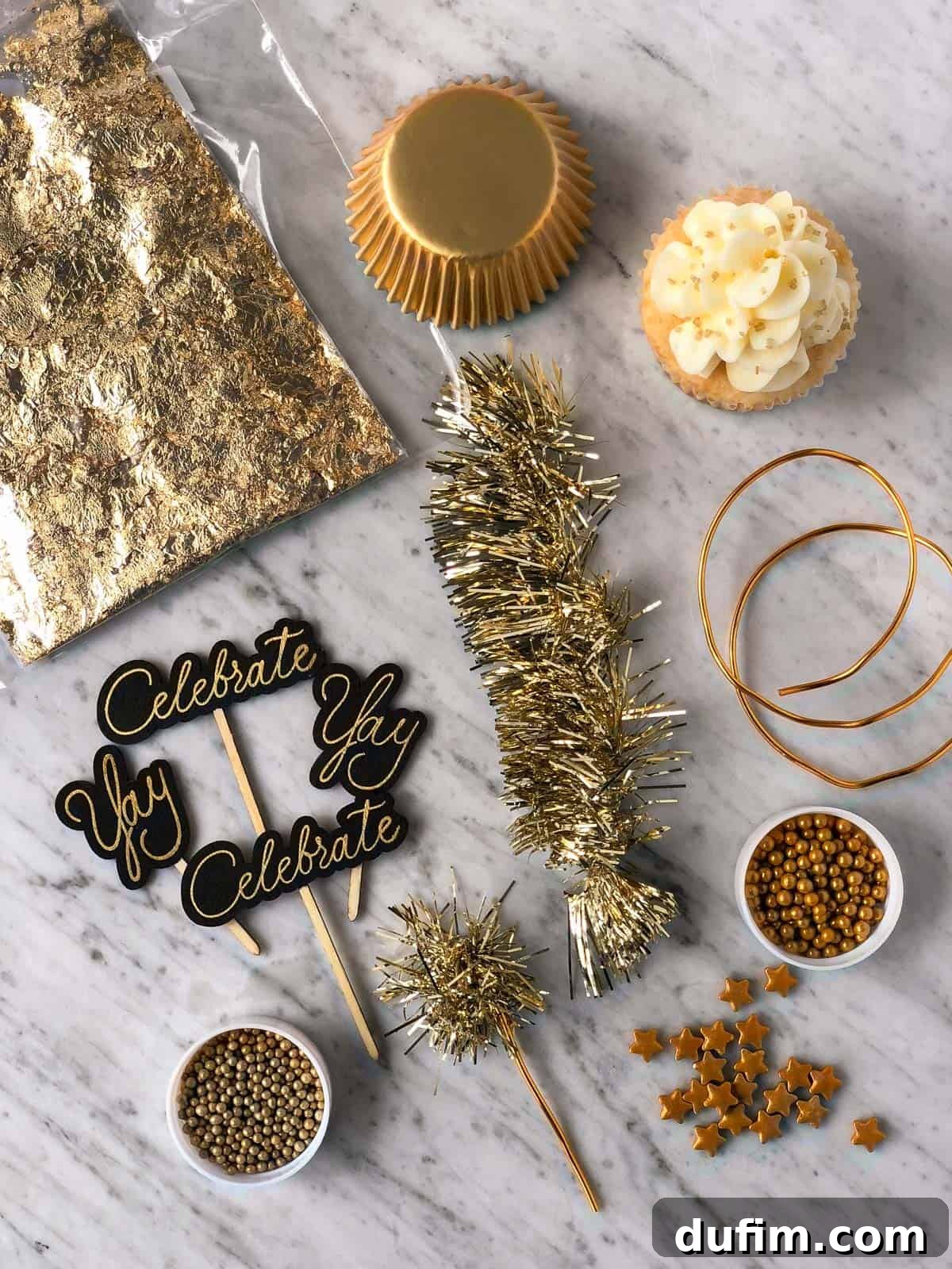 A collection of New Year's Eve cupcake decorating supplies including gold sprinkles, star-shaped sprinkles, gold muffin liners, edible gold leaf, 'Celebrate' and 'Yay' cupcake toppers, and gold tinsel.