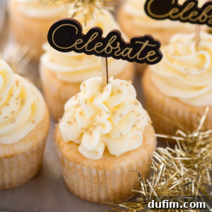 Champagne cupcakes with swirls of white frosting, topped with gold sprinkles and a toothpick sign that says “Celebrate.” Gold tinsel decorations are placed around the cupcakes.