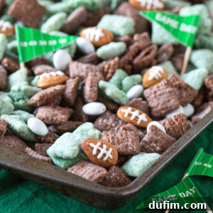 A tray filled with green and brown Chex mix, football puppy chow, white oval candies, football-shaped snacks, and small paper flags that read Game Day, all arranged on a dark green surface.