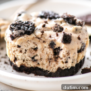 A close-up of a creamy cookies and cream cheesecake topped with crushed chocolate cookies, sitting on a white plate. Its thick cookie crust and partially scooped texture are reminiscent of decadent oreo cheesecake bites.