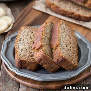 Two thick slices of healthier banana bread sit on a gray plate atop a wooden surface, with more banana bread and fresh banana slices visible in the background.