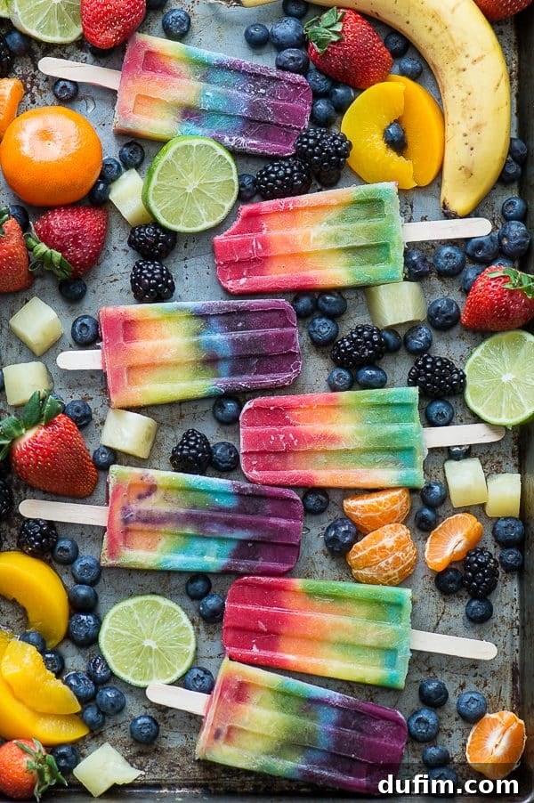 Colorful Rainbow Popsicles surrounded by an assortment of fresh fruits including limes, oranges, blueberries, raspberries, peaches, and bananas.