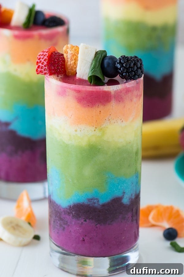 A refreshing Rainbow Smoothie in a clear glass, garnished with fresh blueberries, raspberries, oranges, and bananas.