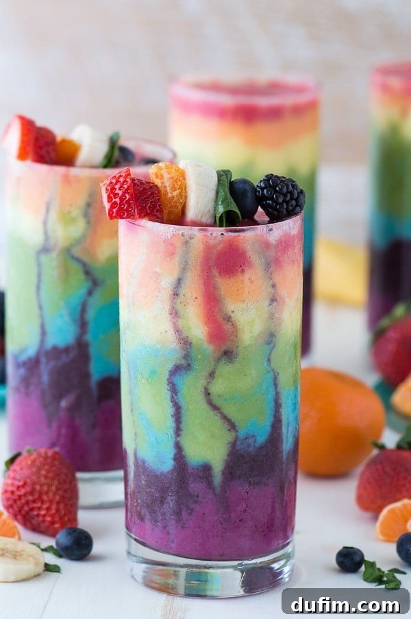 Beautiful 7 layer rainbow smoothie recipe! Full of tons of fruit and topped with a fruit skewer, it’s the ultimate rainbow smoothie! A close-up of a perfectly layered rainbow smoothie in a glass cup, artfully topped with fresh fruit, ready to be enjoyed.