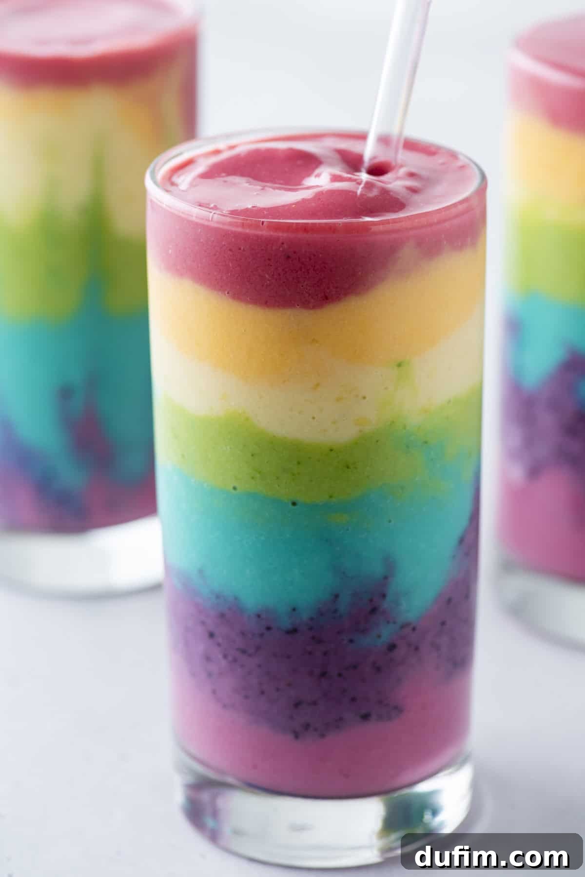 Beautiful 7 layer rainbow smoothie recipe! Full of tons of fruit and topped with a fruit skewer, it’s the ultimate rainbow smoothie! A single, perfectly layered rainbow smoothie in a clear glass cup, resting on a clean white background, showcasing its vibrant colors.