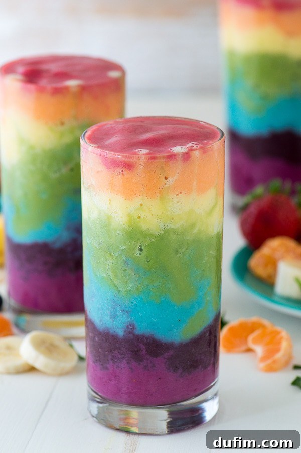 Beautiful 7 layer rainbow smoothie recipe! Full of tons of fruit and topped with a fruit skewer, it’s the ultimate rainbow smoothie! A stunning seven-layer rainbow smoothie, beautifully presented in a glass cup, illustrating the vibrant gradient of colors from bottom to top.