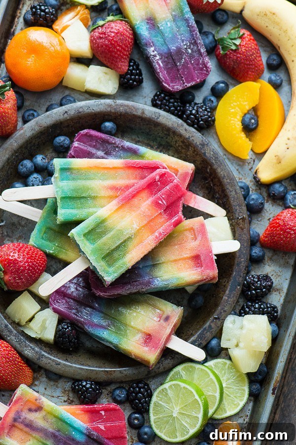 A close-up shot of several rainbow popsicles resting on a metal plate, beautifully garnished with an assortment of fresh berries and citrus slices, highlighting their refreshing appeal.