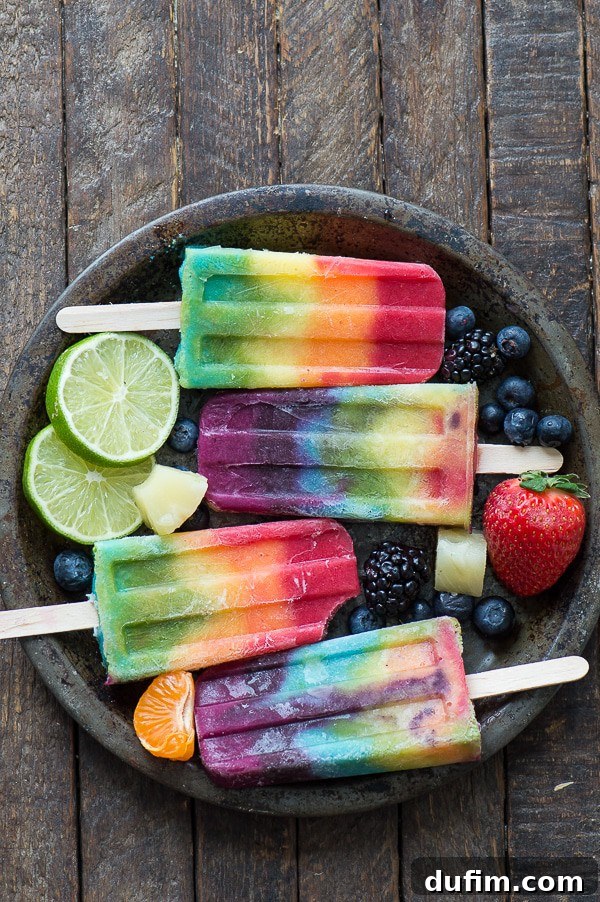 A collection of rainbow popsicles presented on a metal tray, surrounded by vibrant fresh fruits like blueberries, limes, strawberries, and oranges, emphasizing the natural ingredients and colorful layers.