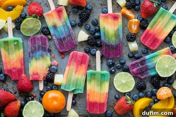 An assortment of colorful rainbow popsicles arranged on a platter, surrounded by fresh and sliced fruits such as blueberries, bananas, oranges, limes, and strawberries, emphasizing their natural goodness.