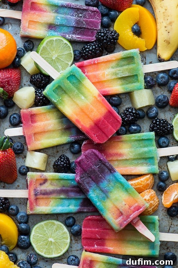 A vibrant display of rainbow popsicles beautifully arranged on a platter, surrounded by fresh and sliced blueberries, bananas, oranges, limes, and strawberries, ready to be enjoyed.