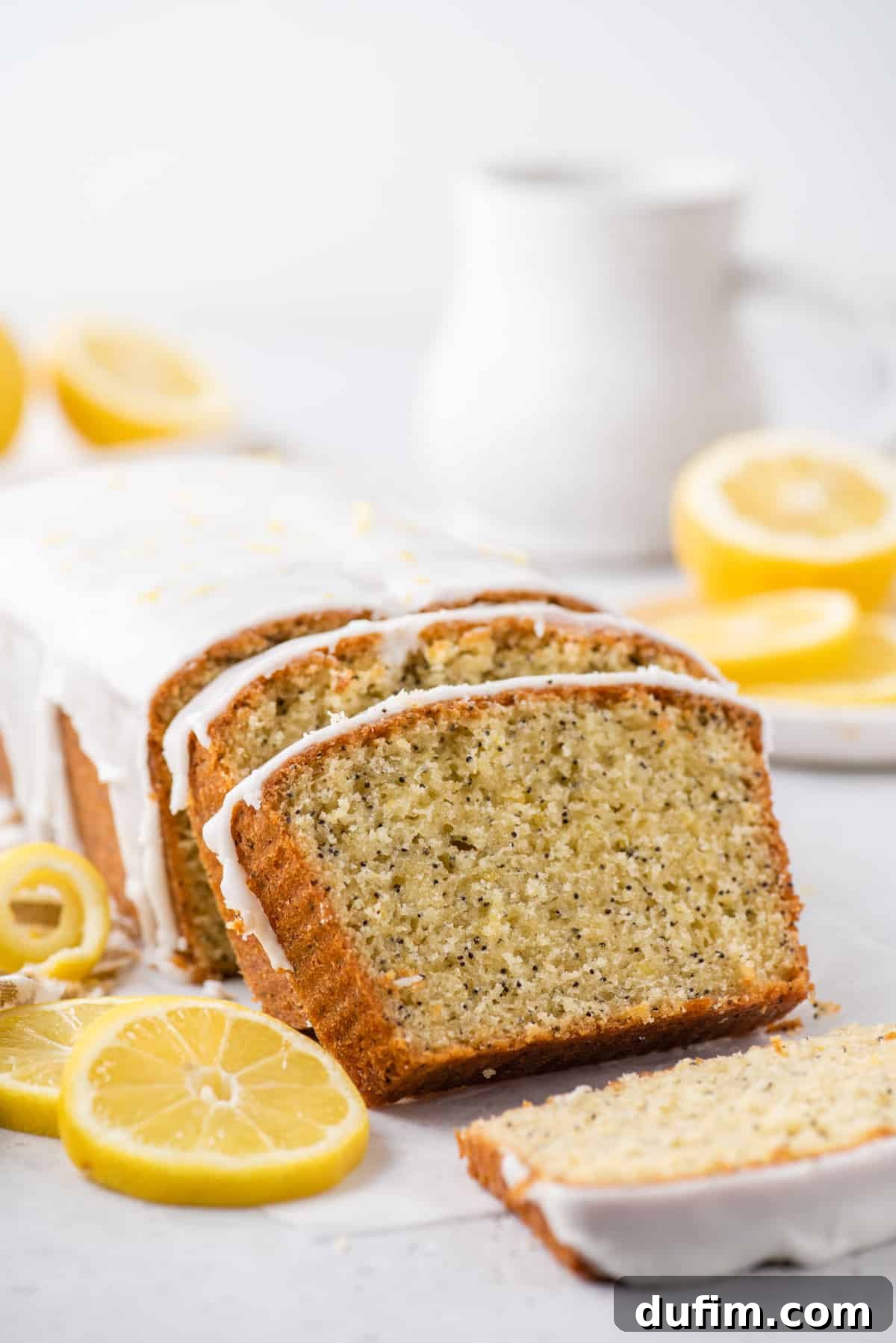 Sliced lemon poppy seed bread with fresh lemon slices and a white pitcher in the background, highlighting its moist texture and vibrant flavor.