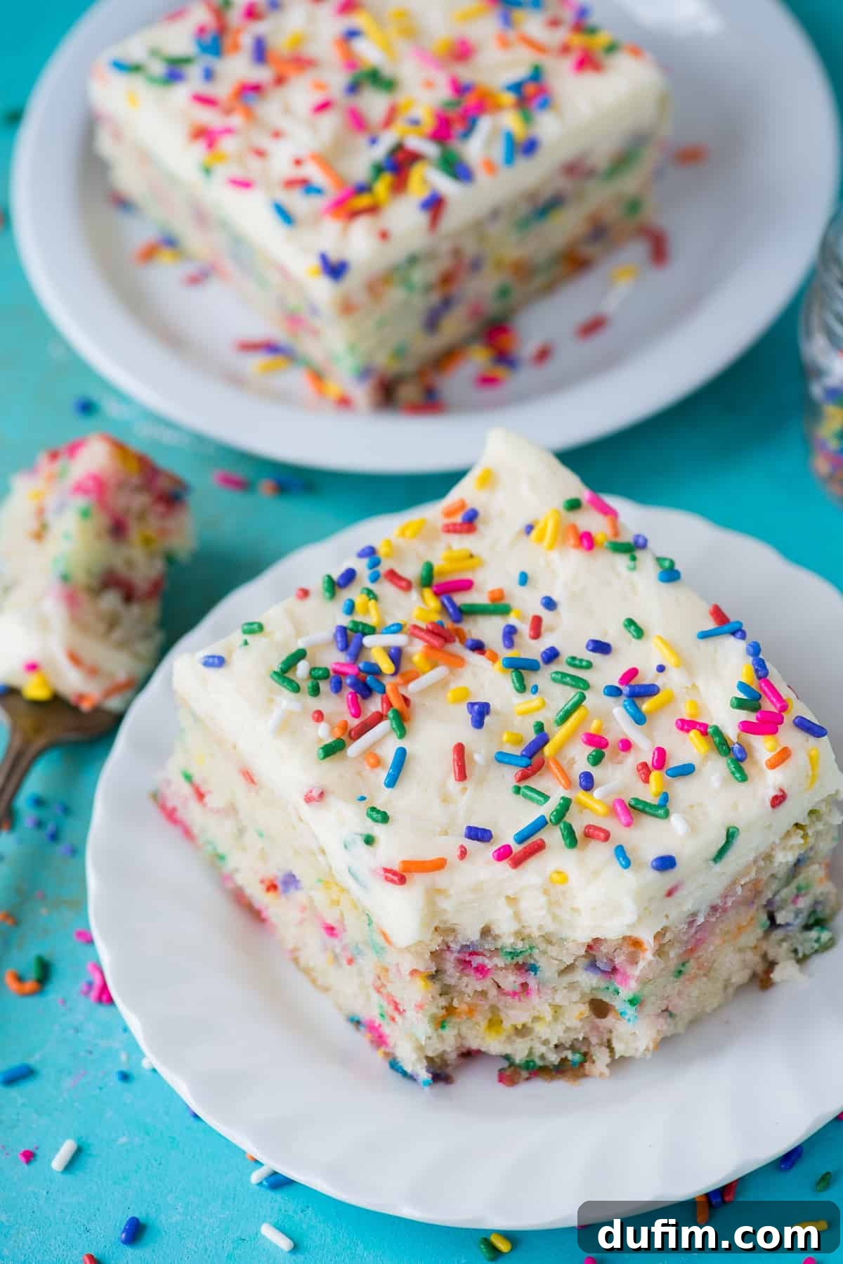 Two generous slices of homemade funfetti cake, adorned with colorful sprinkles and creamy vanilla frosting, elegantly presented on white plates against a vibrant teal background. This image captures the joyful essence of a perfect celebration cake.