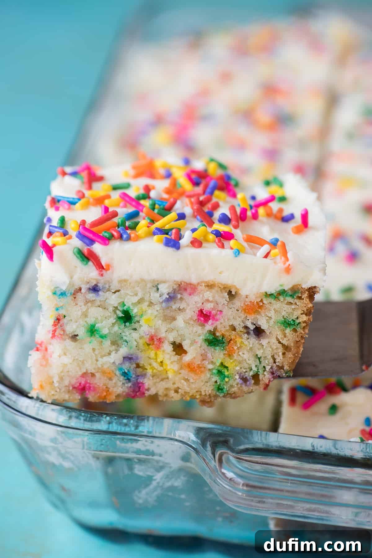 A perfectly baked slice of homemade funfetti cake, complete with colorful sprinkles throughout the cake and topped with smooth vanilla frosting, being lifted on a metal spatula from a baking pan.