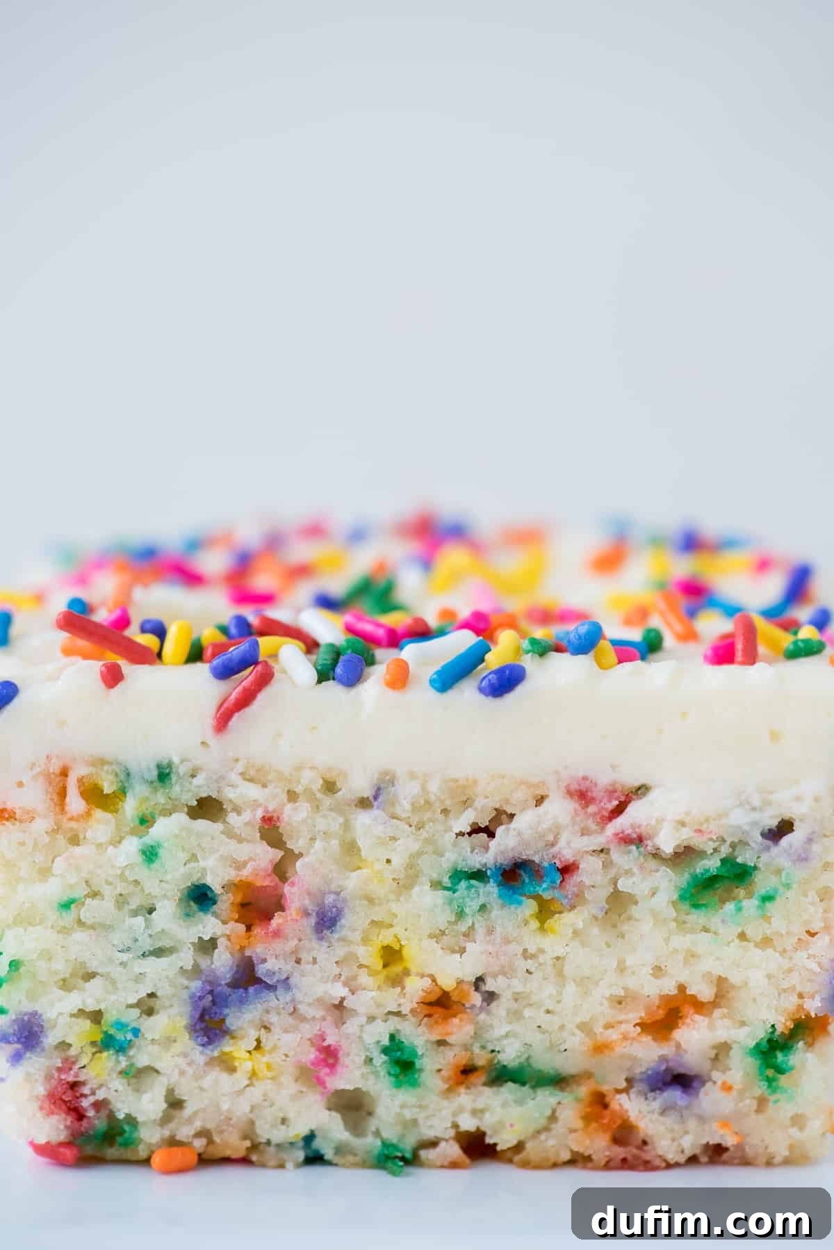 A close-up shot of a single slice of funfetti cake, showing the moist crumb packed with colorful jimmie sprinkles and a thick layer of creamy white vanilla frosting on top.
