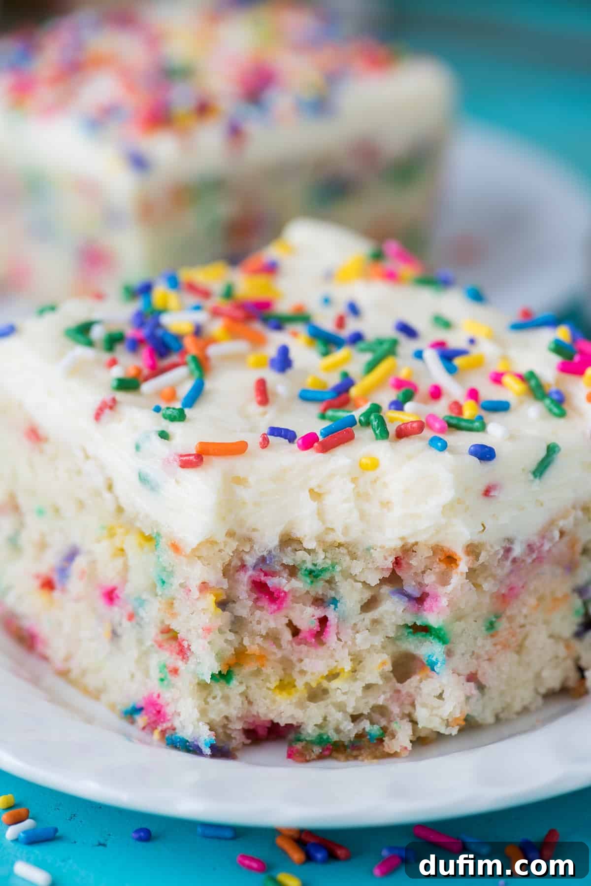 A delectable slice of funfetti cake with a bite taken out, showcasing its fluffy texture and abundance of sprinkles, resting on a white plate against a teal background.