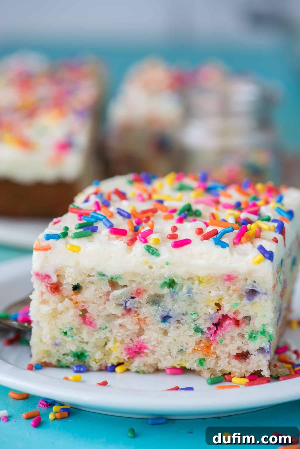 A beautiful, large slice of funfetti cake with white frosting and colorful sprinkles, served on a plain white plate against a serene teal background.