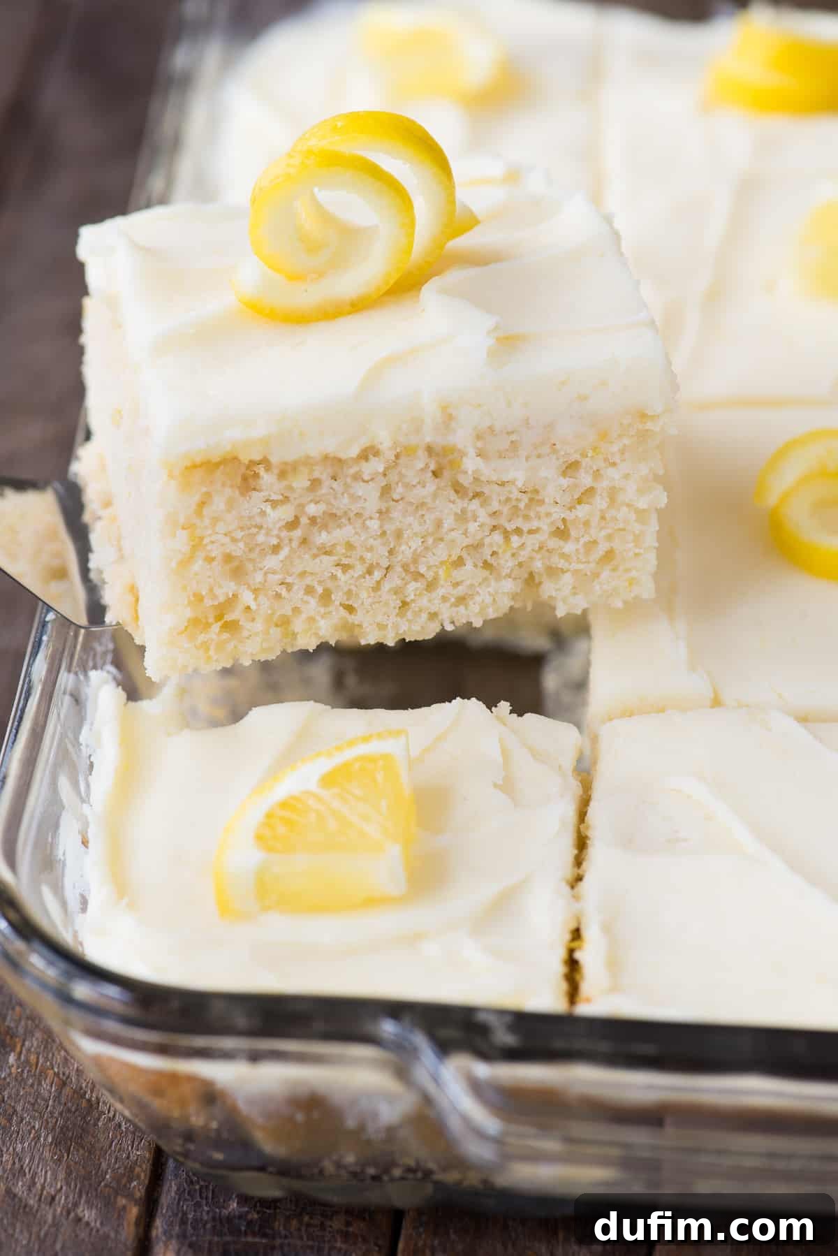 A delectable piece of lemon cake, topped with a luscious white frosting and delicate lemon peel, presented elegantly on a metal spatula.