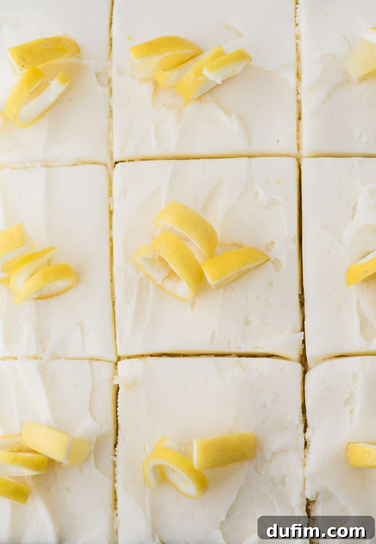 A freshly baked lemon cake, generously covered in white frosting and adorned with thinly sliced lemon peels, cut into perfect serving pieces.