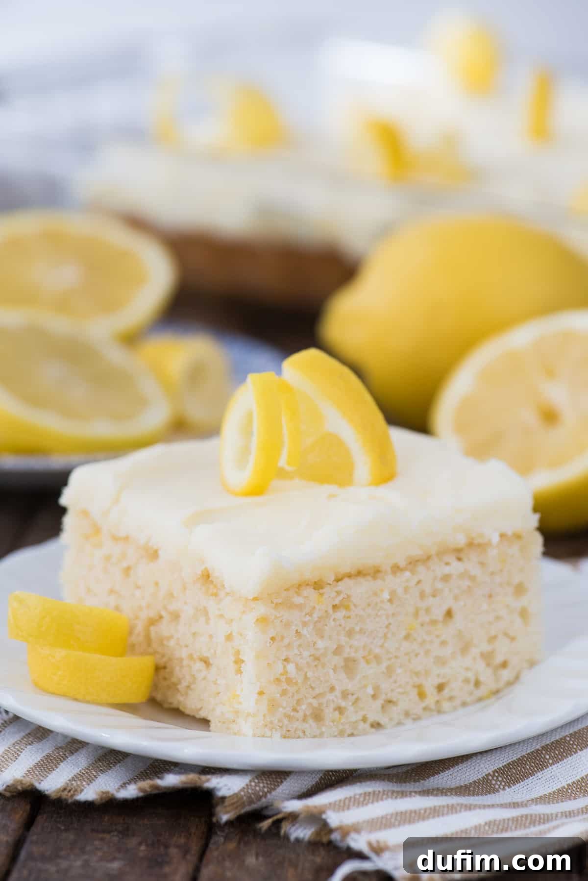 A square piece of lemon cake with a generous layer of lemon frosting, beautifully presented on a white plate with fresh lemons artfully blurred in the background.