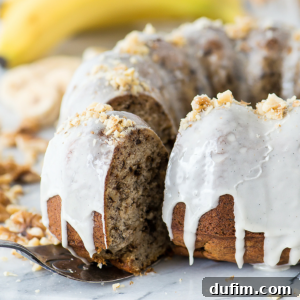 A moist banana nut bundt cake with white icing drips and chopped walnuts on top, presented on a white stand. A slice has been removed, revealing the cake's soft texture. Blurred bananas and walnuts are in the background.