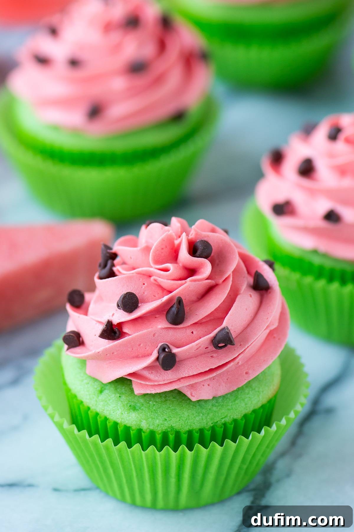 Watermelon Burst Cupcakes 2 Green cupcakes with pink watermelon frosting, adorned with mini chocolate chips resembling seeds, presented on a elegant marble background.