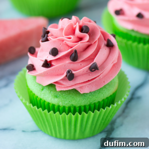 A vibrant watermelon cupcake with a green cake base and pink swirled frosting, topped with mini chocolate chips, set in a green liner on a marble background.