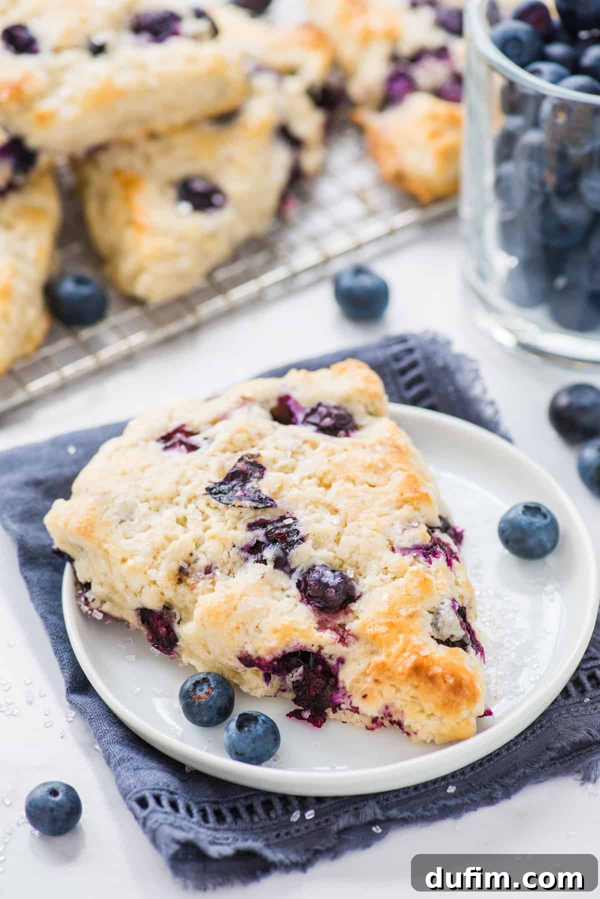 Tender Blueberry Buttermilk Scones 2 A perfectly baked blueberry scone resting on a white plate with a blue napkin, showcasing its golden crust and tender interior.