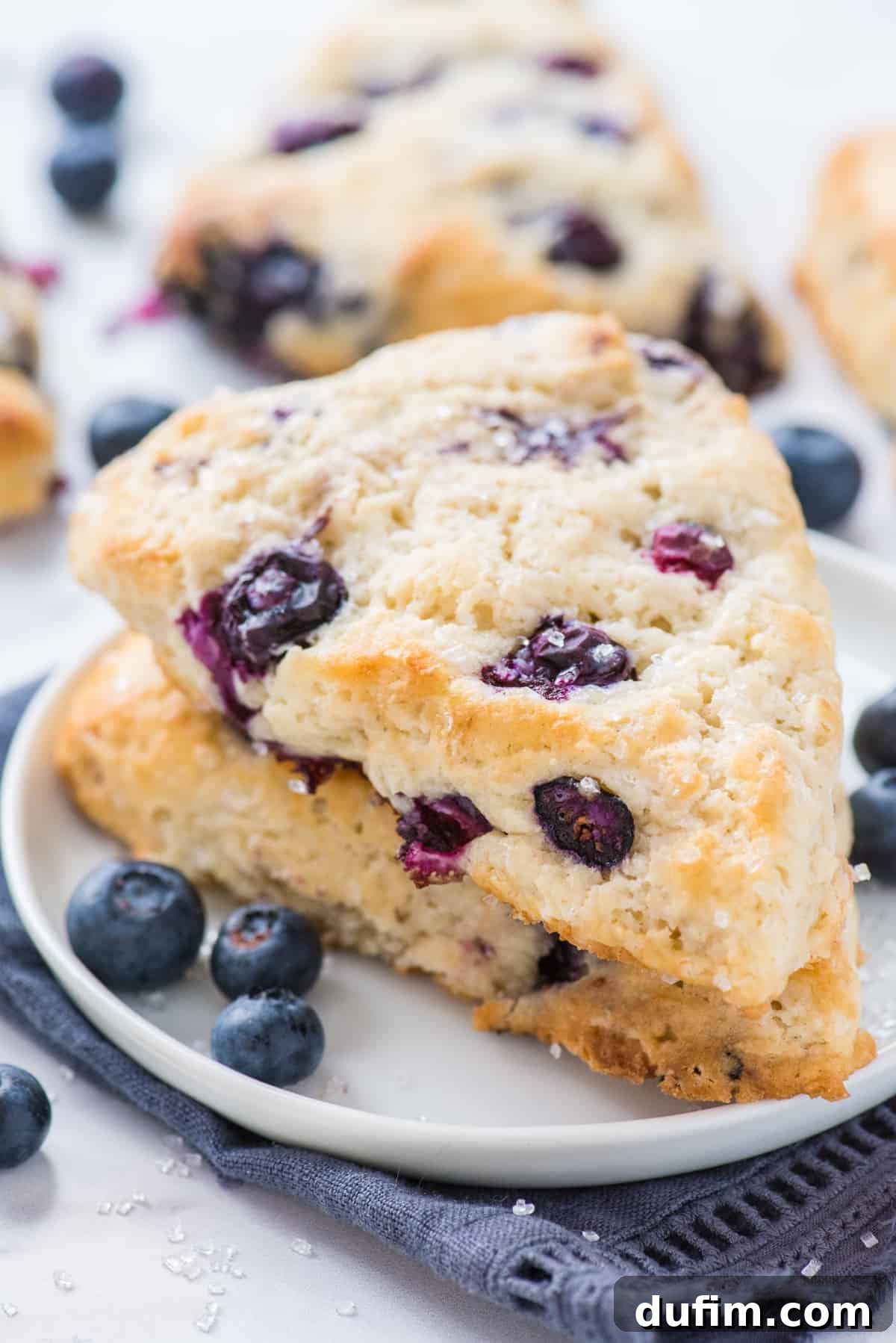 Tender Blueberry Buttermilk Scones 13 Two freshly baked blueberry scones stacked on a white plate, accompanied by a blue napkin, highlighting their golden crusts and tender layers.
