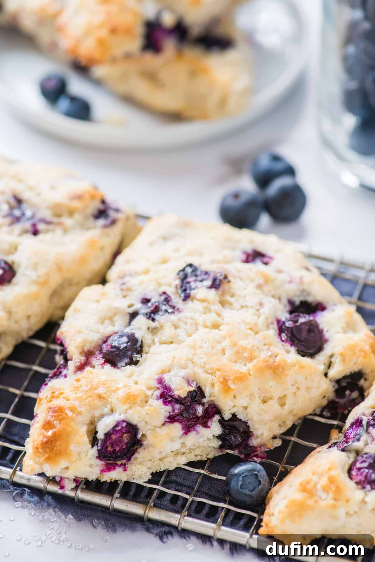 Tender Blueberry Buttermilk Scones 14 Freshly baked blueberry scones cooling on a wire rack, with a blue napkin underneath, set against a clean white background.