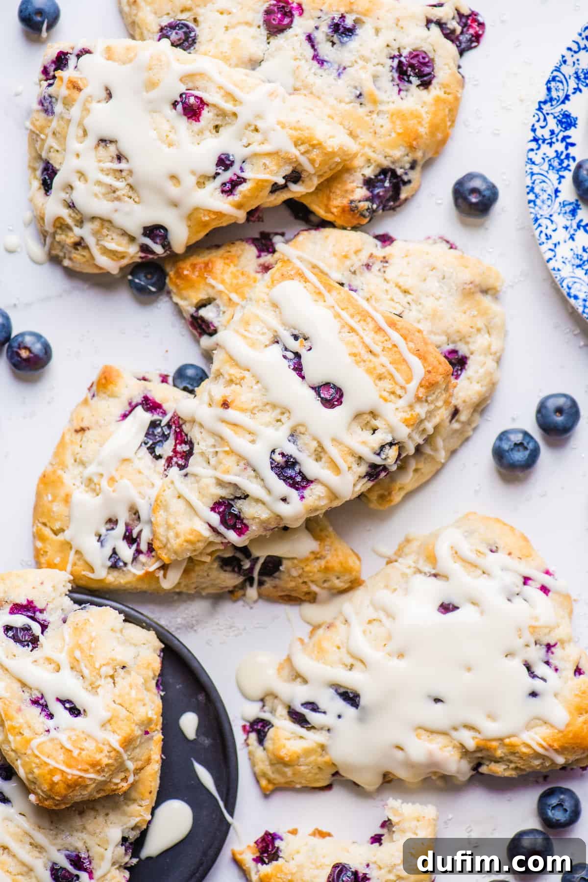 Tender Blueberry Buttermilk Scones 3 Several blueberry scones, beautifully glazed with a light white icing, are artfully arranged on a clean white background, ready to be enjoyed.