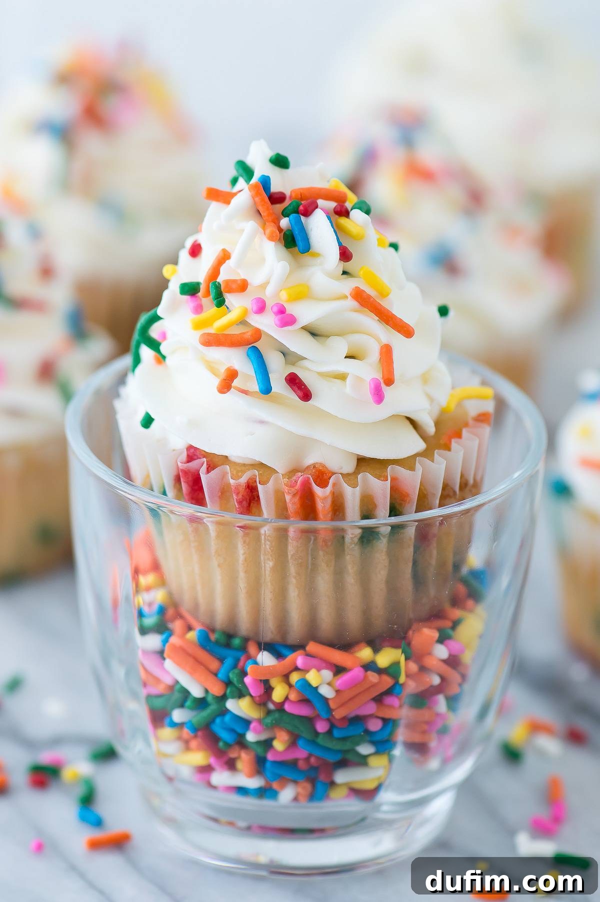 A delicious funfetti cupcake in a clear glass, showing off its sprinkle-filled interior, surrounded by loose sprinkles.