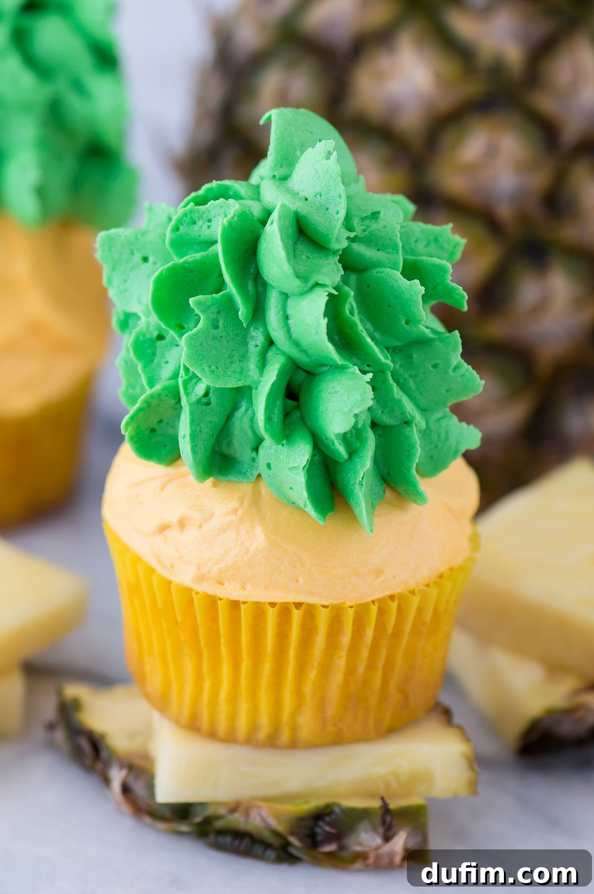 A close-up of a single pineapple cupcake being decorated, showing the piping bag adding green leaves to the yellow buttercream base.