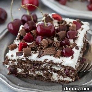 A square slice of black forest icebox cake with layers of chocolate and cream, topped with fresh cherries and chocolate shavings, served on a gray plate with a fork.
