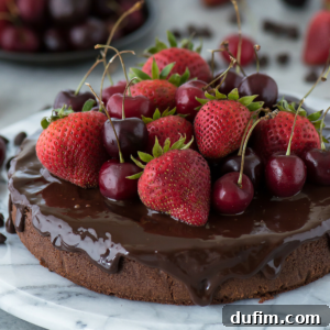 A rich mud cake topped with glossy chocolate ganache, fresh strawberries, and cherries sits on a marble surface with more fruit in the background.