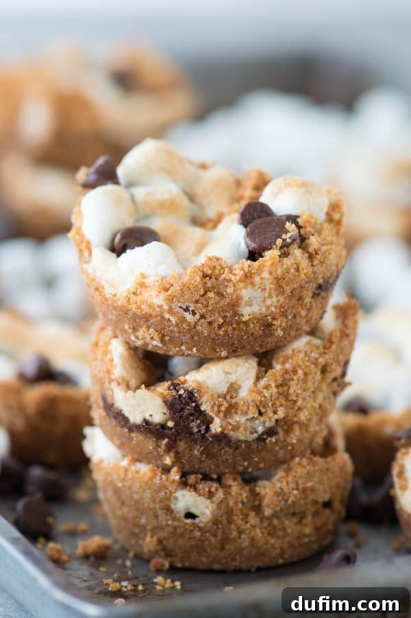 Three perfectly stacked easy S’mores Cups, highlighting their distinct layers of graham cracker, chocolate, and toasted marshmallow, surrounded by more delectable cups on a baking sheet.