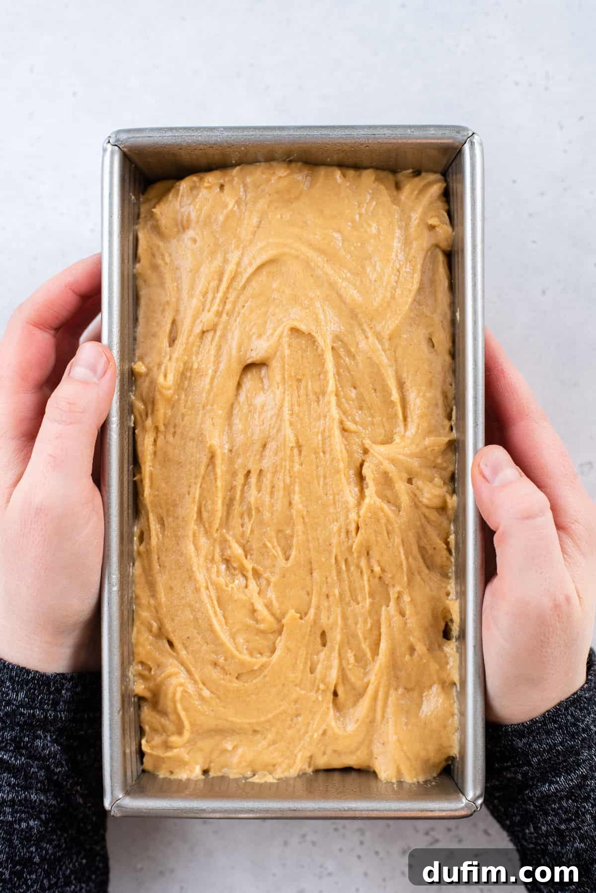 Hands holding a metal loaf pan filled with peanut butter bread batter before baking